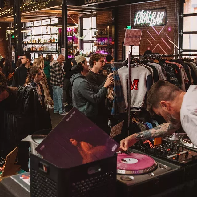 photo of a dj at a turntable at a vintage & vinyl event. people drink beverages and peruse racks of clothes